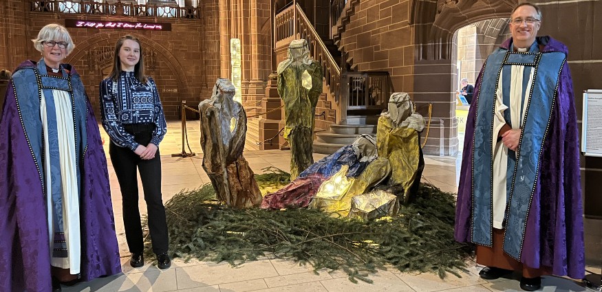 A male vicar and a female vicar and a young female student stand next to a nativity scene.